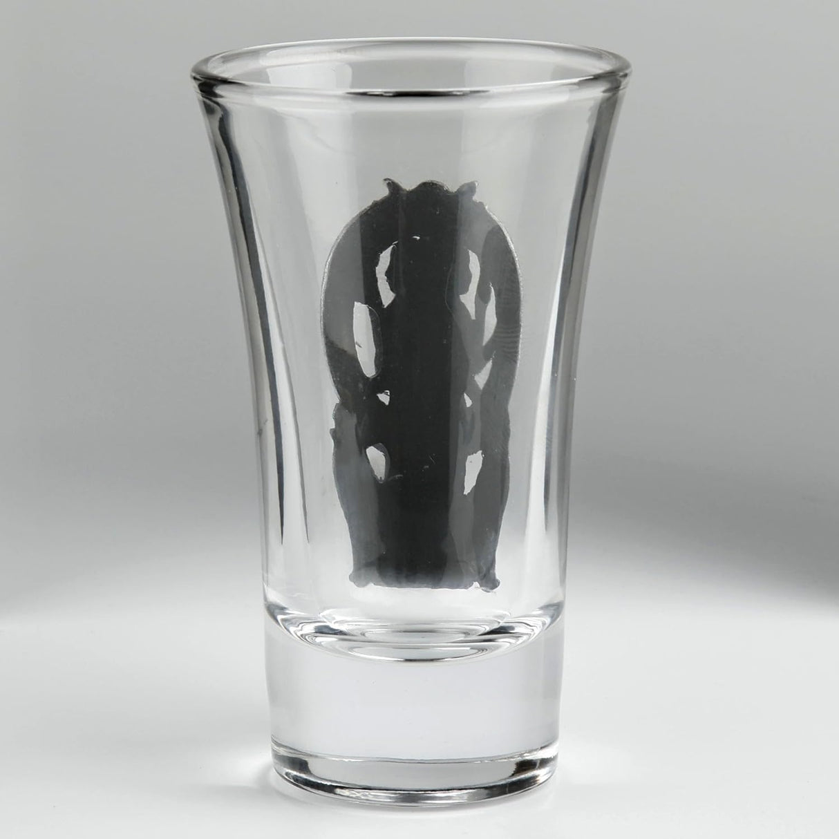 A clear glass shot glass featuring a detailed pewter relief of a triple moon goddess figure, Hecate, flanked by two wolves. Shown from the back