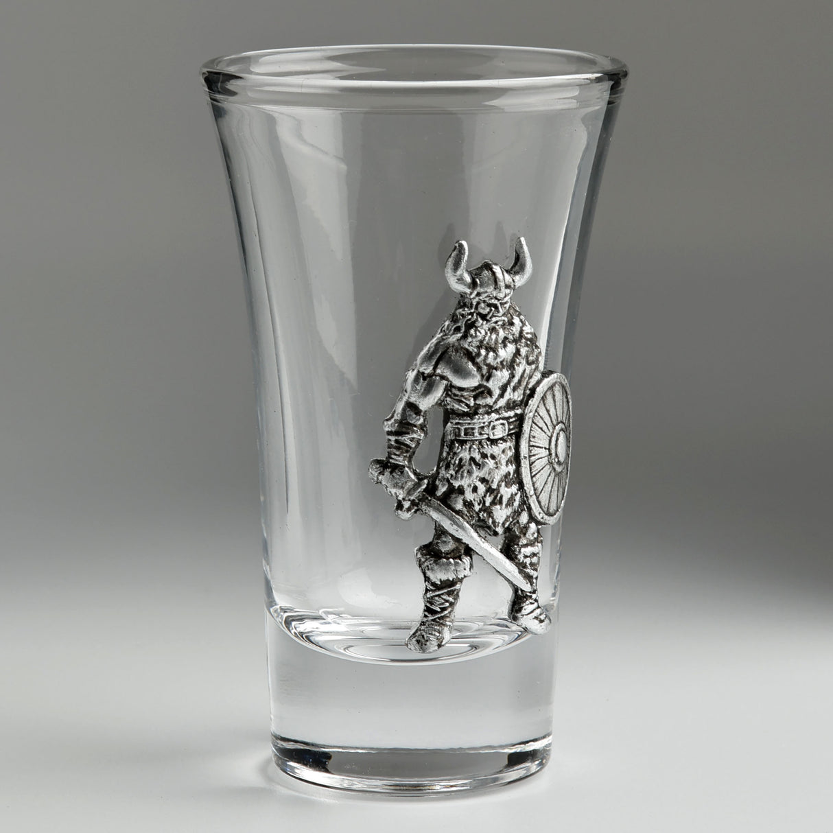 A clear glass shot glass featuring an embossed pewter Viking warrior holding a shield and sword