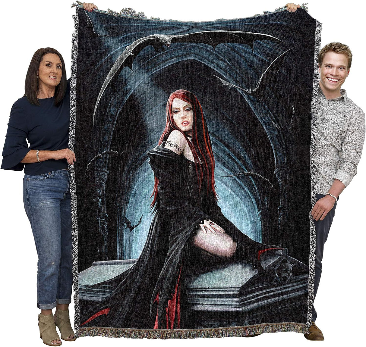 A large woven throw blanket with fringe edges is held up by a man and a woman against a plain white background. The blanket features a gothic illustration of a vampire woman with long red hair, surrounded by bats in a dark blue and black archway scene.