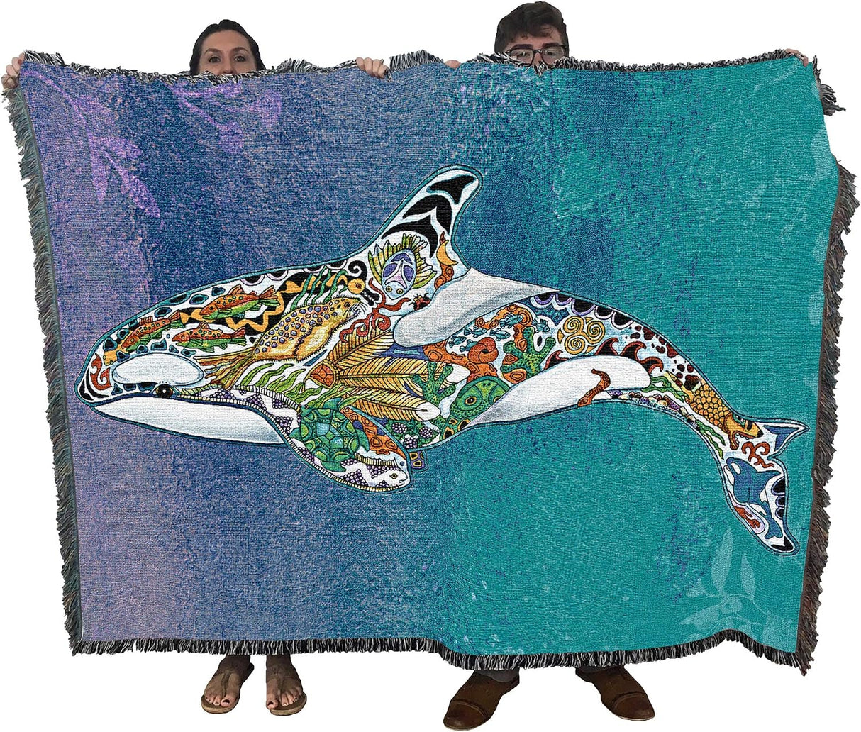 A large woven throw blanket with fringe edges is held up by two people against a white background. The blanket features a colorful, patterned orca whale design over a blue and teal gradient background.