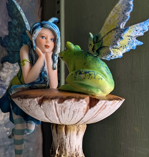 A figurine featuring a fairy with blue wings conversing with a green winged frog perched on a mushroom.