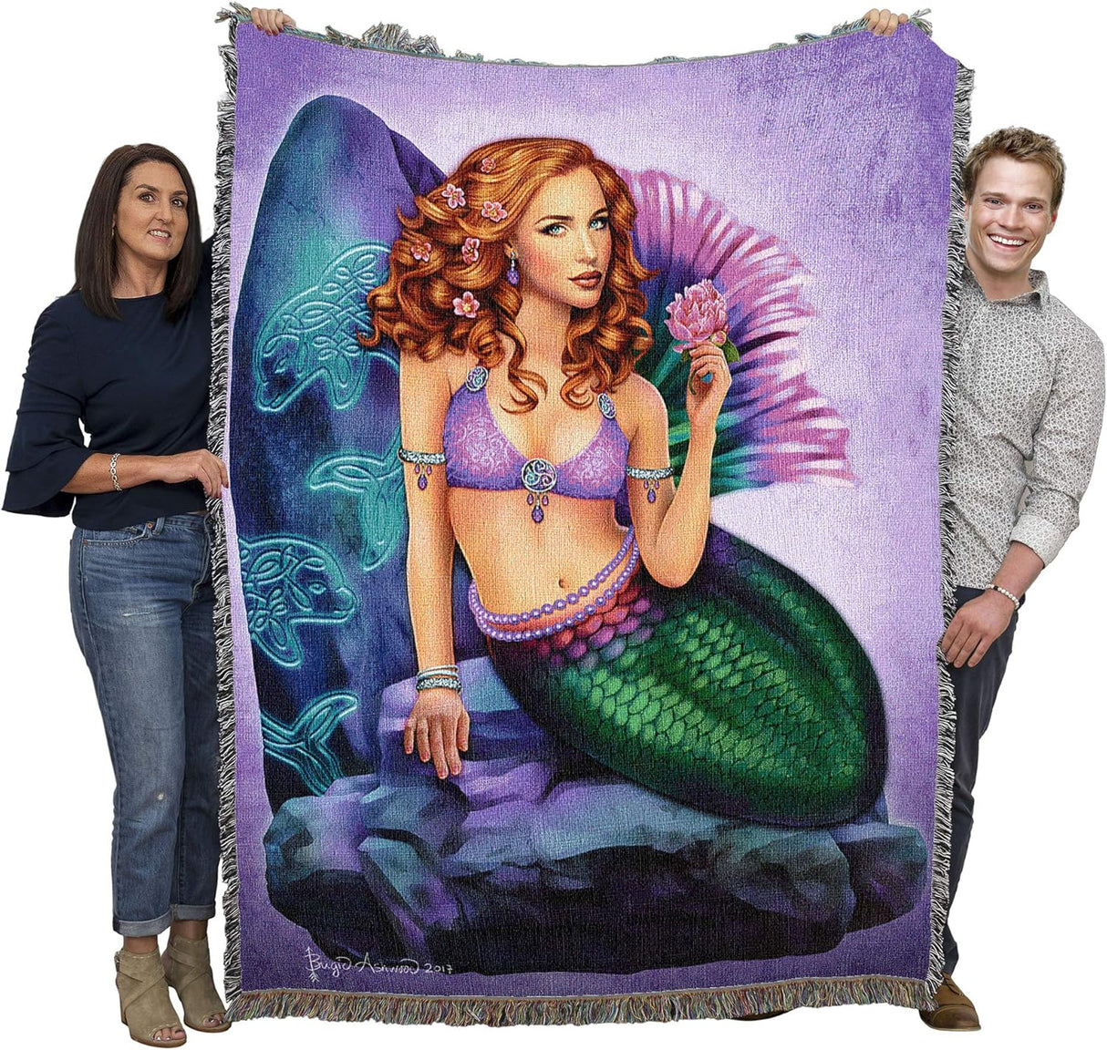 Tapestry blanket, art by Brigid Ashwood, mermaid with mostly green tail, fins in purple and pink. Red hair, holding pink flower, sitting against stone carved with Celtic knotwork dolphins. Blanket shown held up by two adults to show large size