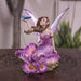 Fairy figurine; she has brown hair and a purple dress surrounded by matching flowers. She looks up at the blue butterfly perched on her outstretched hand. Shown on a wooden table
