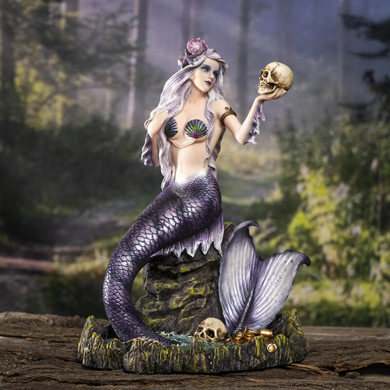 Mermaid holding up a skull figurine. Her scales are black and purple and she wears an iridescent seashell bra. Her hair is light violet with a seahsell. She sits on a rock with her tail in a pool of water, with another skull and golden treasures. Shown on a forest backdrop