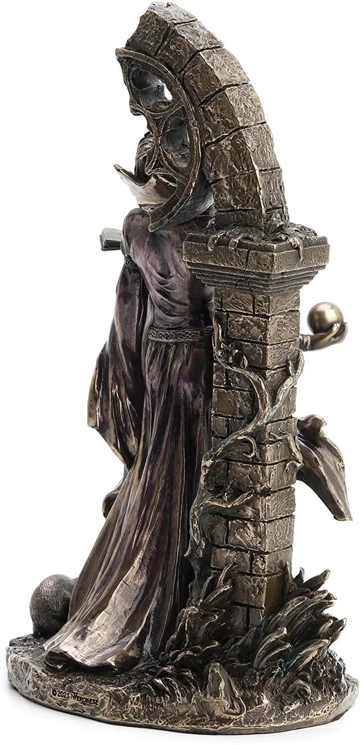 Side view - Aradia s represented standing with eyes closed, face tipped to the heavens. Her dress is long and flowy, and she holds a tome in one hand and an orb in the other. She stands in front of ruins twined with vining flowers.