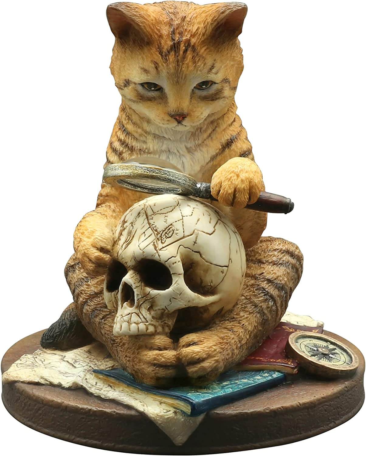 Figurine of an orange tabby cat holding a magnifying glass up to a human skull. The kitty sits amidst books, maps and a compass.