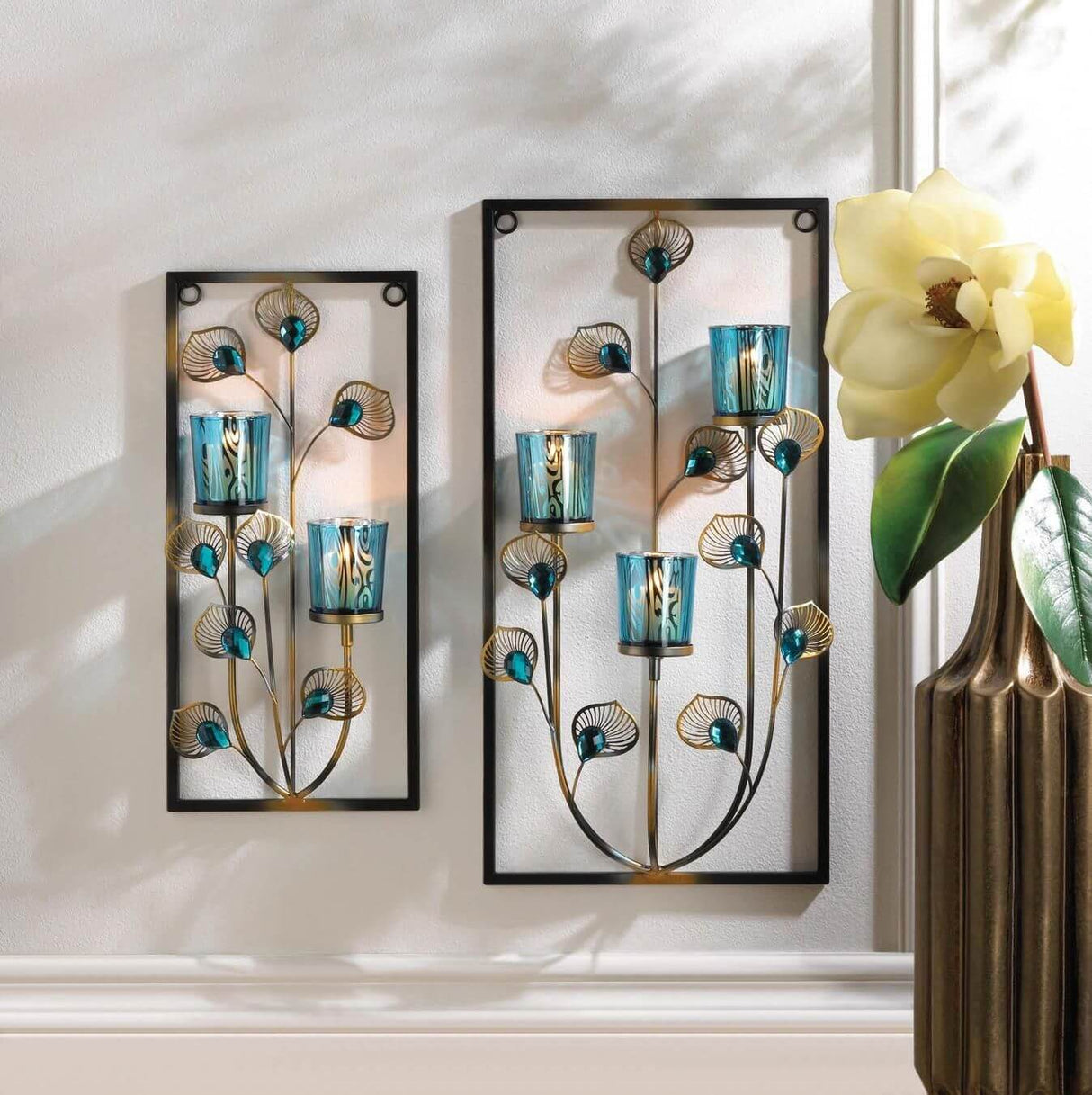 Peacock themed wall sconce candle holder. Votive holders in shades of blue are held amidst jeweled feather-like stems in a rectangular frame. A two candle holder and three candle holder version are displayed next to each other, hung on a sunny wall near a flower.