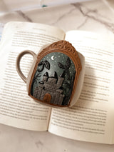 Decorative mug with a castle and dragon design in clay, displayed on an open book