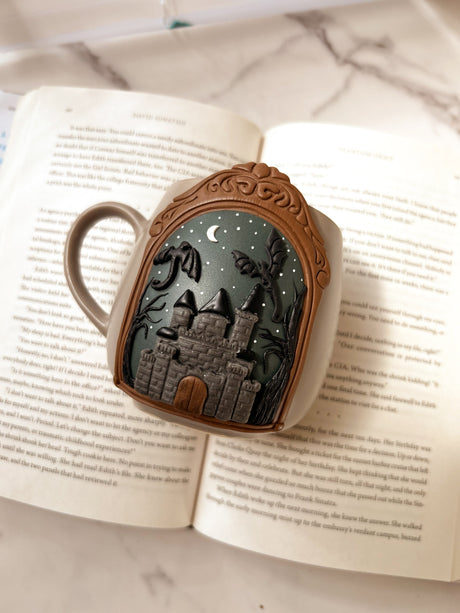 Decorative mug with a castle and dragon design in clay, displayed on an open book