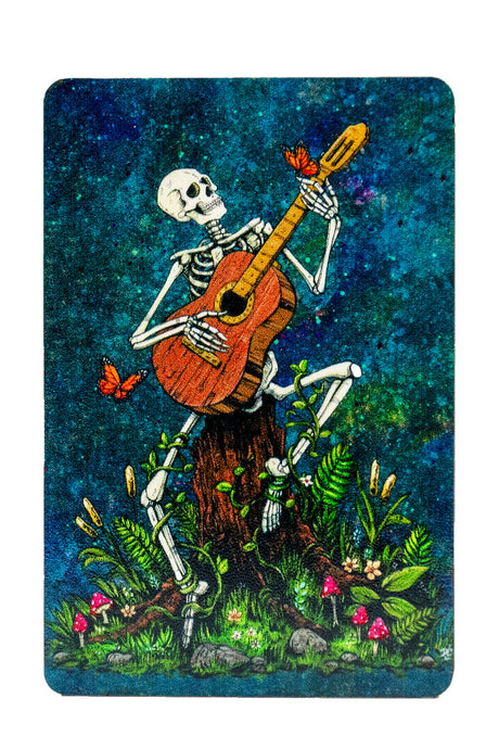 Magnet showing a skeleton playing acoustic guitar with butterflies, surrounded by flowers and vines, sitting on a tree stump. Blue mottled background.