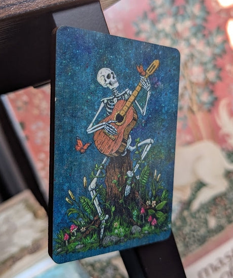 Magnet showing a skeleton playing acoustic guitar with butterflies, surrounded by flowers and vines, sitting on a tree stump. Blue mottled background.