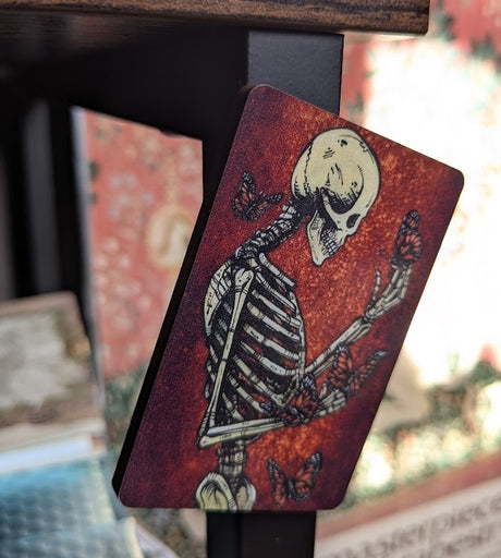 Magnet of a skeleton contemplating and surrounded by monarch butterflies on a red background