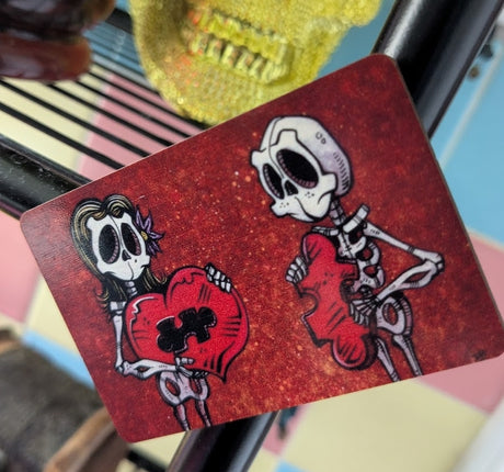 Magnet with two skeletons, one holding a heart with missing section and the other holding the puzzle piece that fits inside