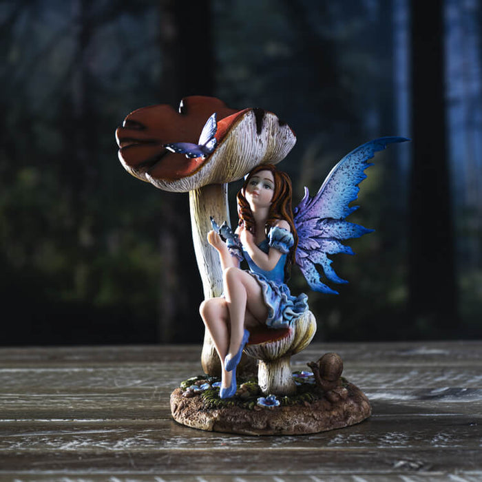 The statue shows a fairy sitting on a toadstool. She wears a dress of ruffled blue, and has indigo wings to match. A butterfly perches on her hand, and another purple flutterby sits on a taller mushroom. 