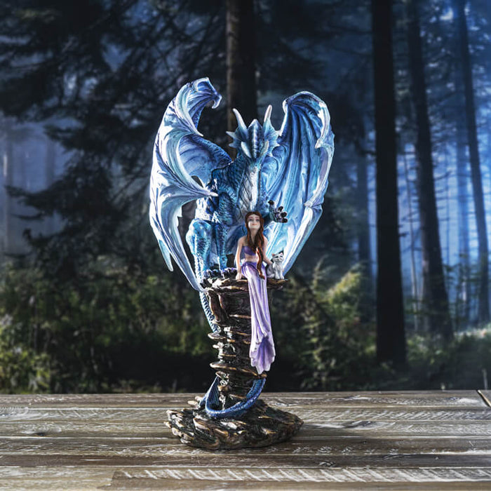 Blue dragon protectively curling wings around fairy in purple and white cat. Shown in a forest setting on a wood table
