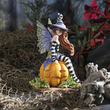 Figurine of a fairy in black and white stripes with purple and green accents sitting on a pumpkin next to a black cat