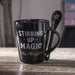 Coffee mug in black with "STIRRING UP MAGIC" text and stars, spoon slots into handle and has a pentacle