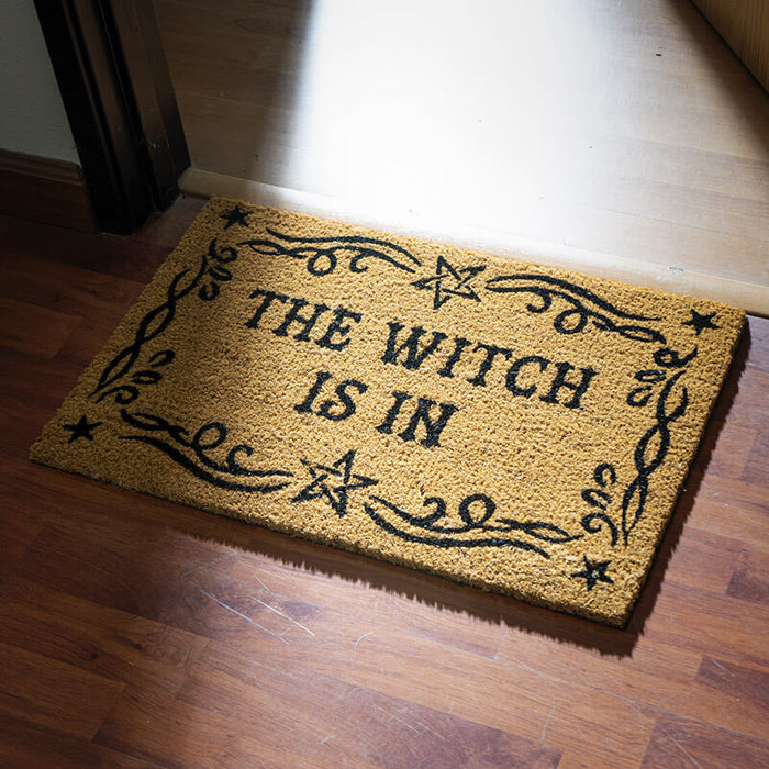 Natural tan doormat with black text reading "THE WITCH IS IN" surrounded by a star and swirl border
