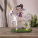 Figurine of Betty Boop with fairy wings and purple flowers in her hair, riding a unicorn in a grassy meadow, shown on a table