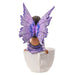 Figurine of a fairy with purple wings and dress and dark skin sitting on the edge of a teacup, back view