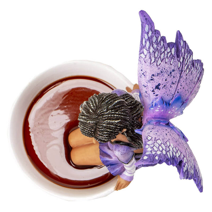 Figurine of a fairy with purple wings and dress and dark skin sitting on the edge of a teacup, top down view
