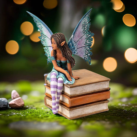 Figurine of a tanned skin fairy with brown braided hair and teal-purple wings in a blue-green dress, perched on a stack of tan books. Striped white and purple stockings.