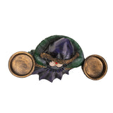 Decorative figurine of a witchy woman in a green cloak and purple dress holding two bowls into which candles can sit, shown top down