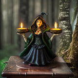 Decorative figurine of a witchy woman in a green cloak and purple dress holding two bowls into which candles can sit, shown with candles lit