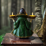 Decorative figurine of a witchy woman in a green cloak and purple dress holding two bowls into which candles can sit, shown with candles lit from the back