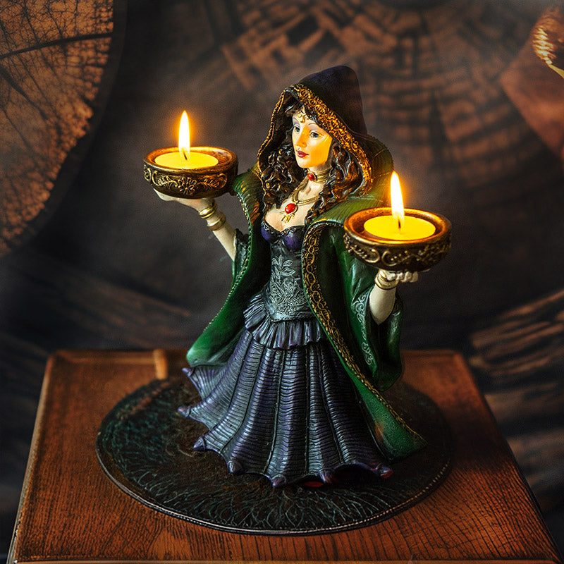 Decorative figurine of a witchy woman in a green cloak and purple dress holding two bowls into which candles can sit, shown with candles lit