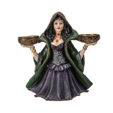 Decorative figurine of a witchy woman in a green cloak and purple dress holding two bowls into which candles can sit