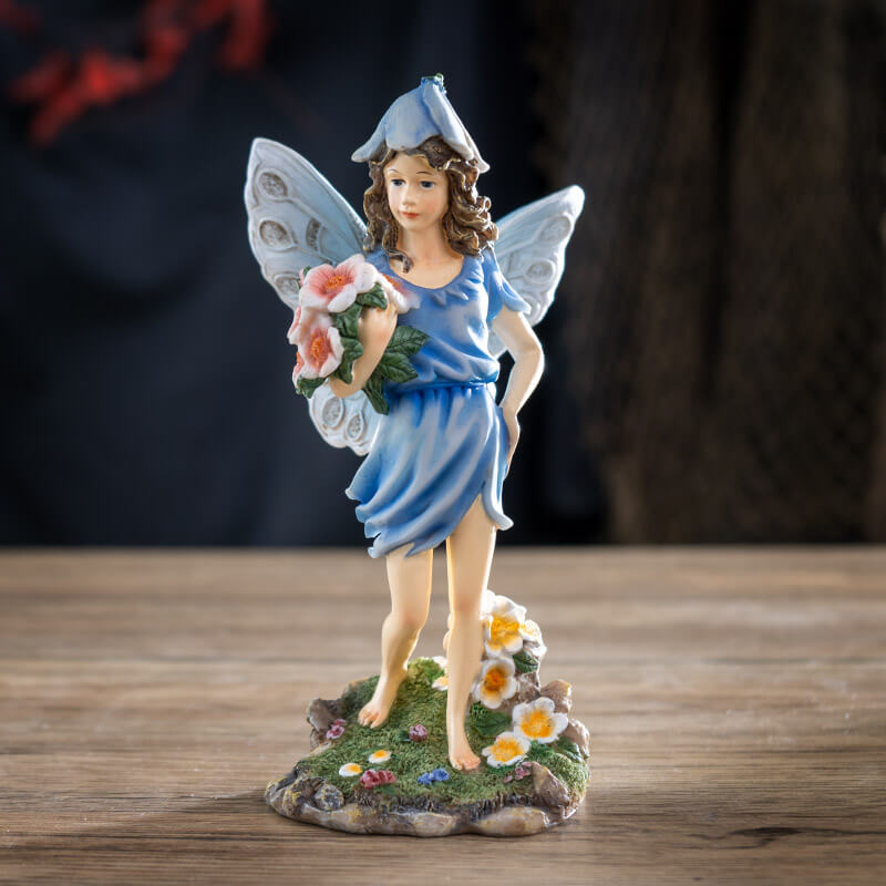 Figurine of fairy in a blue dress with flower hat and pale wings holding bouquet of pink flowers with more blooms at her feet.