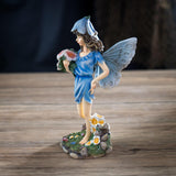 Figurine of fairy in a blue dress with flower hat and pale wings holding bouquet of pink flowers with more blooms at her feet.