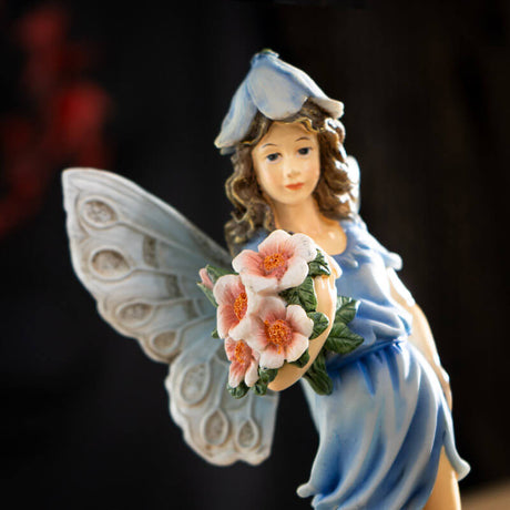 Figurine of fairy in a blue dress with flower hat and pale wings holding bouquet of pink flowers with more blooms at her feet.