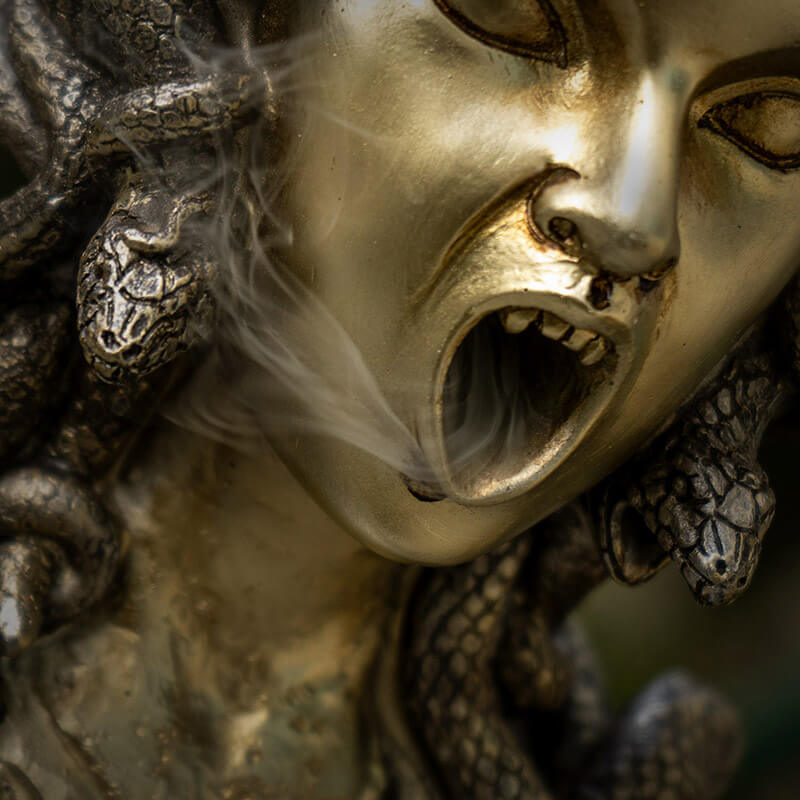 Backflow incense burner shaped like medusa's face with open mouth for smoke, snake hair. Done in faux-bronze. Closeup of mouth with smoke