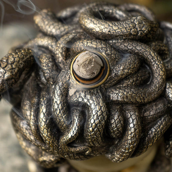 Backflow incense burner shaped like medusa's face with open mouth for smoke, snake hair. Done in faux-bronze. Shown with incense.