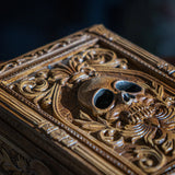 Tarot or trinket box in brown hue with skull and elaborate designs on lid, more fleur de lis emblems on the side. Closeup of lid, resembling faux-wood