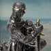 faux-metallic figurine of a medieval knight kneeling, holding helm and sword, wearing chainmail with gold accents