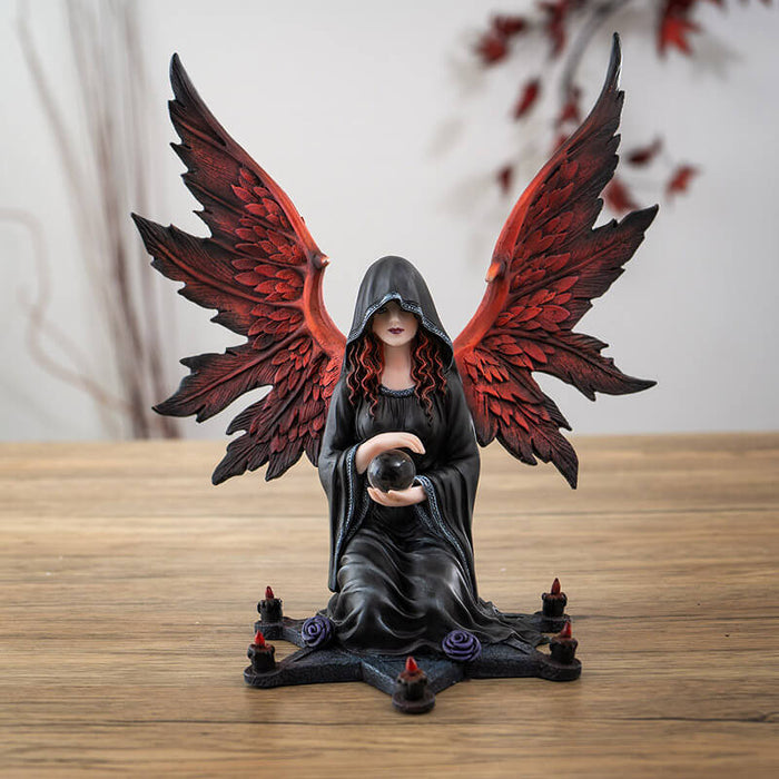 Figurine of an angel with red wings and hair in a black cloak, holding a crystal ball sitting at the center of a star with purple roses and candles