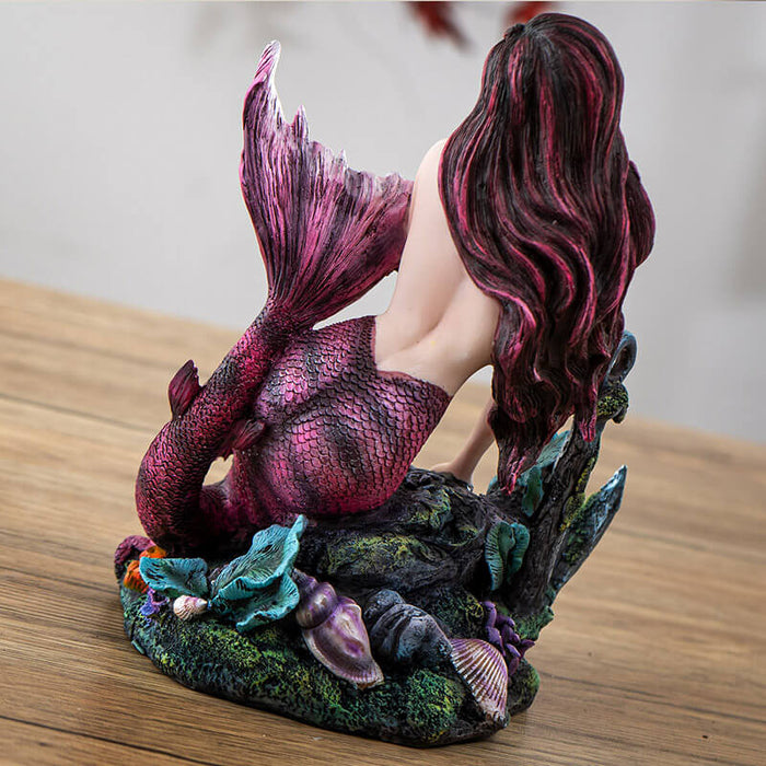 Figurine of mermaid with pink hair and magenta and black striped tail sitting with an anchor, fish, skull and coral, shown from the back
