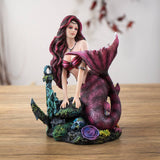 Figurine of mermaid with pink hair and magenta and black striped tail sitting with an anchor, fish, skull and coral