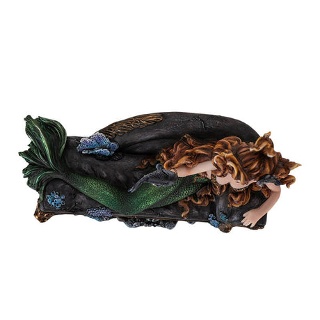 Figurine of a mermaid reclining on a black couch. She has green scales and auburn hair and holds a fish on one hand.