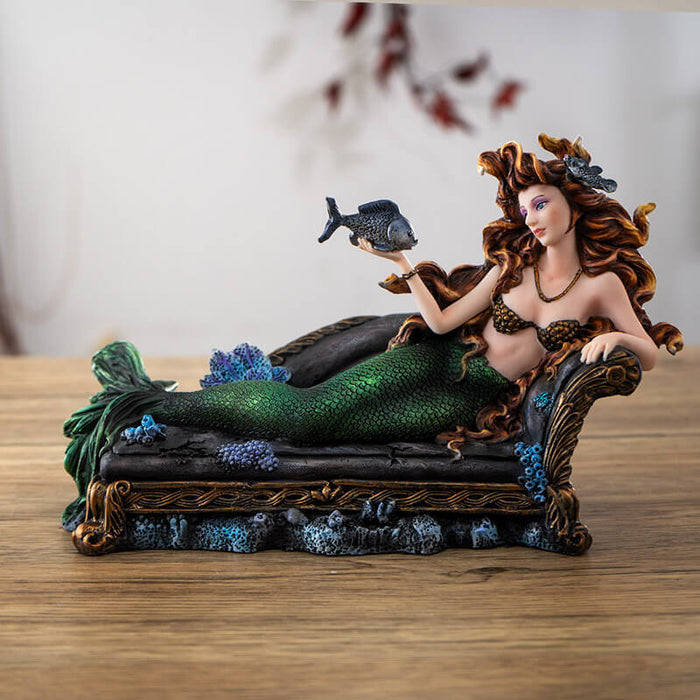Figurine of a mermaid reclining on a black couch. She has green scales and auburn hair and holds a fish on one hand.