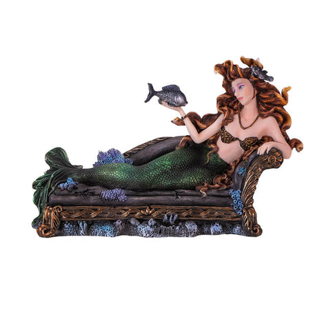 Figurine of a mermaid reclining on a black couch. She has green scales and auburn hair and holds a fish on one hand.