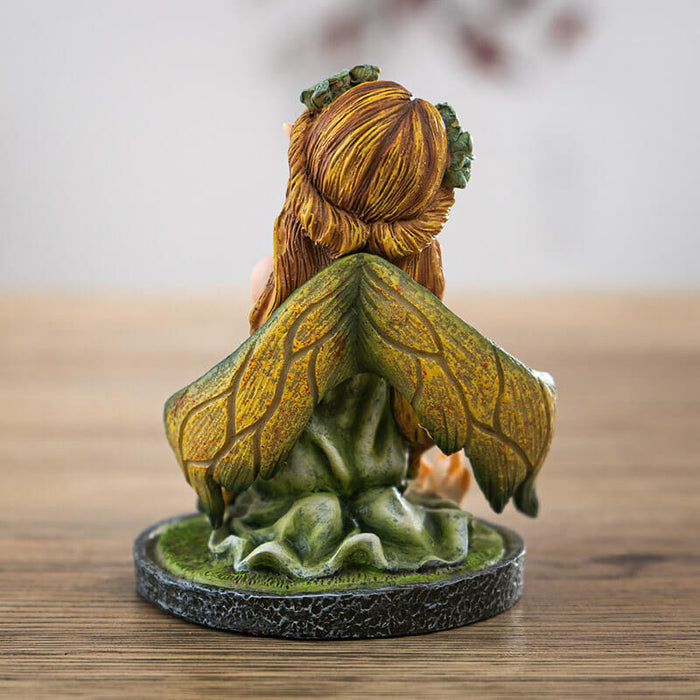 Figurine of small fairy with crystals and mushrooms where she kneels in the moss, holding a bouquet of flowers with more in her blond hair. Shown from the back