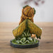Figurine of small fairy with crystals and mushrooms where she kneels in the moss, holding a bouquet of flowers with more in her blond hair. Shown from the back