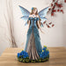 Figurine of a fairy in a blue dress with detailed blue wings and long hair, standing with bright indigo flowers.