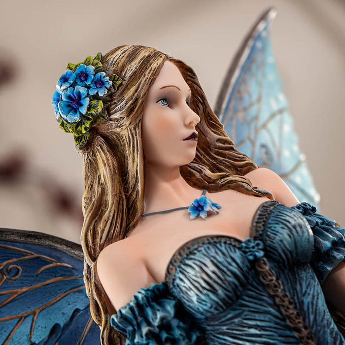 Figurine of a fairy in a blue dress with detailed blue wings and long hair, standing with bright indigo flowers.