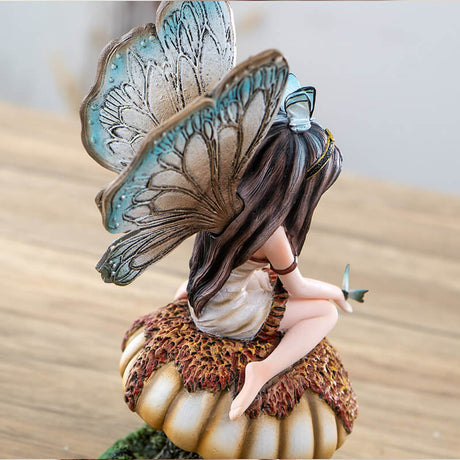 Figurine of brunette fairy with tan and brown wings and cream dress sitting on mushroom with butterfly on her hand, more toadstools below.