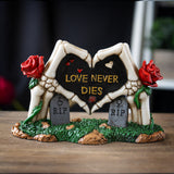 Figurine of skeleton hands forming heart with roses and gravestones, text reading "LOVE NEVER DIES"